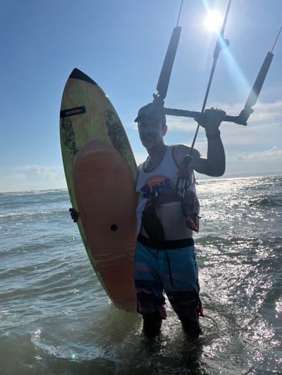 This is a vertically oriented, close-up photograph taken from a low angle, showing a person engaged in kitesurfing. The subject, seen from the waist up, is holding onto a control bar with both hands, gripping it with gloved hands. They are wearing a white sleeveless shirt with a colorful graphic design that includes orange and yellow circular elements and the word "ONE" in orange letters. The lower half of the subject's body is clad in blue, patterned boardshorts. They are standing on a surfboard, the front of which is prominently visible, angled towards the left and the sky. The surfboard has a camouflage-like pattern in shades of green and yellow, with a black tip and a darker section near the tail. The word "f-one" is visible in black text on a white rectangle on the surfboard.
The scene is set on the ocean, with choppy water glistening under bright sunlight. The sun itself is a prominent feature, appearing as a lens flare in the upper left quadrant of the image, casting a strong light that creates a radiant effect on the water's surface. The sky is a bright, clear blue with a few wispy clouds. The overall impression is one of an active, sunny day at the beach, likely during midday given the strong sunlight and clear skies. The image captures the dynamic nature of kitesurfing, with the board cutting through the water and the control bar ready for action.
