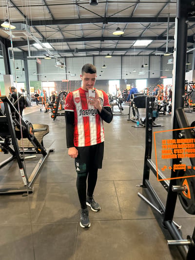 The image displays a full-length mirror selfie of a young adult male inside a Basic-Fit fitness facility, located in Hœnheim, France. The male subject stands centrally, presenting short dark hair, a light mustache, and a goatee. He is dressed in a red and white vertically striped short-sleeved athletic jersey, worn over a black long-sleeved undershirt. Black athletic shorts are worn over black compression leggings, with a green knee sleeve visible on his right leg. His footwear consists of dark athletic shoes with white soles. He holds a white smartphone horizontally with both hands, angled towards the mirror, and exhibits a slight smile. A ring is present on his left ring finger.
The gym floor is constructed of dark gray rubber mats. The facility features a high, open industrial-style ceiling with exposed steel trusses, ductwork, skylights, and numerous suspended light fixtures. Various black and orange Basic-Fit branded fitness equipment is distributed throughout the spacious area. In the background, multiple other individuals are discernible, engaged in diverse exercise routines. To the right of the main subject, a black weight rack system displays an orange translucent sign with French text: "LE SEUL MOMENT OÙ TU PEUX BAISSER LES BRAS, C'EST POUR RANGER TES POIDS" (The only time you can lower your arms is to put away your weights), alongside an illustrative graphic depicting a weight plate and a storage rack.