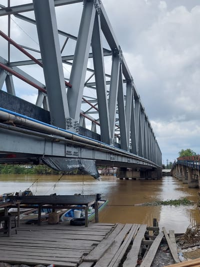 This is a media file taken in Parit Baru, Indonesia. The primary subject is a large, gray metal truss bridge extending over a muddy brown river. The bridge's intricate metalwork is prominent in the foreground, with visible bolts and supporting beams. Along the side of the bridge, there are blue and grey pipes. A black tarp with white text is also attached to the side of the bridge structure.
The river flows next to the bridge, reflecting the overcast sky. On the right bank of the river, there is a wooden dock with several small boats moored. Some of these boats appear to be traditional fishing vessels. Various objects are scattered on the dock, including what look like buckets, a tire, and some wooden planks.
In the background, another bridge structure with concrete pillars and railings is visible, lined with trees. The sky is cloudy, suggesting it is daytime, likely overcast or nearing dusk. The overall impression is of a functional, perhaps somewhat industrial, riverine environment.
The text visible on the black tarp reads: "PEMBANGUNAN JEMBATAN Tingkat Kabupaten Sampang".