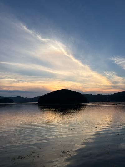 This is a photograph of a sunset over a body of water, likely a lake or river, with silhouetted hills in the background. The sky is a gradient of blue, transitioning to hues of orange and pink near the horizon where the sun is setting. Wispy clouds stretch across the sky, catching the last rays of sunlight. In the foreground, the water is calm, reflecting the colors of the sky. A small, dark, silhouetted island or large rock is visible in the middle ground, with more silhouetted hills and trees forming the distant shoreline. There are a few small boats on the water in the distance. The overall scene evokes a sense of peace and natural beauty. The location is identified as Gródek nad Dunajcem, Poland.