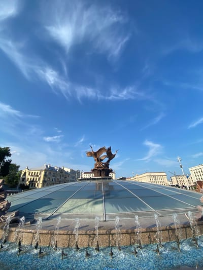 The image captures a bright, sunny day in Minsk, Belarus, likely mid-morning or early afternoon given the clear blue sky with wispy clouds and strong sunlight. The scene is dominated by a modern fountain with a blue tiled basin and a glass enclosure. Water streams from multiple spouts along the edge of the fountain, creating a dynamic visual element.
In the center of the fountain, a bronze sculpture of birds in flight is visible. The birds appear to be swans or cranes, with outstretched wings. Behind the fountain, a row of light-colored, multi-story buildings with visible windows lines a street. The architecture suggests a European city.
The image is oriented vertically, with the sky occupying the majority of the frame. The fountain and buildings are seen from a low angle, emphasizing their scale against the expansive sky. There is no visible text in the image. The overall mood is one of peacefulness and urban beauty on a clear day.