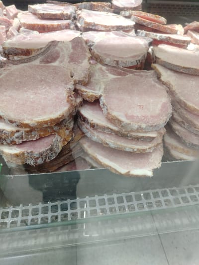 A close-up, eye-level shot shows a display of sliced pork chops in a refrigerated case, likely in a supermarket in Juan Griego, Venezuela. The pork appears to be frozen, with a white frost coating the edges of the cuts, which vary in thickness. The chops are stacked in multiple piles, revealing their pink meat and visible layers of fat. The display case has a metal grate at the bottom, visible through a glass front, and the lighting suggests an indoor commercial setting.