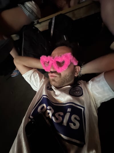 A person wearing pink heart-shaped glasses and a white jersey with blue trim and the word "oasis" printed in large white letters on a blue background is lying down with their arms behind their head. The person has short brown hair and a beard. Their right foot, wearing a blue sandal, is visible. A black phone is tucked into the front of their jersey. The environment appears to be indoors with dark surroundings. The image is taken from a low angle, looking up at the person. The person appears to be in a relaxed or playful pose. There is no indication of the location being in Washington Township, United States of America.