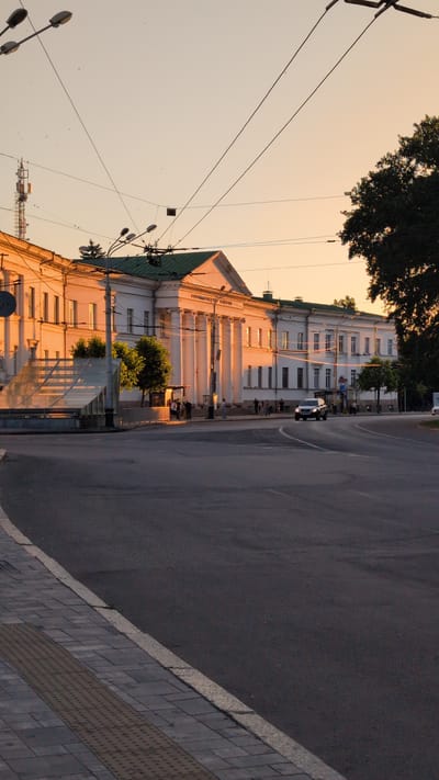 A wide-angle, vertical shot captures a street scene in Poltava, Ukraine, during what appears to be sunset. The golden light of the setting sun bathes a grand white building on the left side of the frame, casting long shadows. This building, featuring a colonnade and a green roof, dominates the background. In the foreground, a wide asphalt road curves towards the right. A pedestrian walkway with grey paving stones and textured tactile paving runs along the left edge of the road. Several streetlights and overhead wires crisscross the sky, with a communication tower visible in the upper left corner. A dark-colored SUV is driving on the road. A few individuals are visible standing near the building and on the walkway, appearing to be pedestrians. Trees line the right side of the street, partially obscuring the building. The sky is a soft gradient of pale orange and yellow.