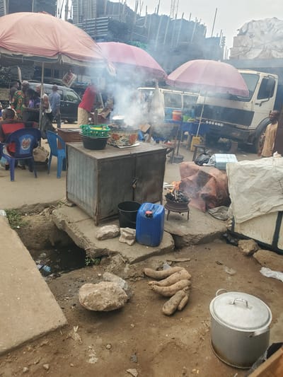 The image depicts an outdoor food stall in what appears to be Fegge, Nigeria. Several individuals are gathered near the stall, seated on blue plastic chairs. A vendor in a red shirt is visible behind a metal table where food is being prepared, with smoke rising from cooking. The stall is shaded by pink umbrellas. A truck is visible in the background. Adjacent to the food preparation area, on the ground, are a large metal pot with a lid, several raw yams, and a stone. A blue plastic container and a black bucket sit on a concrete platform near a small fire in a grill. The ground is dirt and concrete with a drainage ditch. The background shows a metal construction.