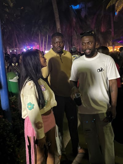 The image captures a nighttime outdoor gathering in Accra, Ghana, illuminated by dim ambient light and various colored stage or event lights in the background. In the foreground, three individuals are centered. On the left, a woman with long dark hair wears a cream-colored cropped cardigan featuring embroidered or crocheted pastel flowers (light purple, light blue, brown) and a short, fringed pink skirt. She holds a smartphone in her left hand and is turned towards the right, engaging with the man opposite her. Behind her, a man with a short beard and dark hair wears a mustard yellow polo shirt, standing with a neutral expression, observing the scene. On the right, a second man with a beard and dark hair, wearing sunglasses pushed onto his head, sports a white t-shirt with a black "OLK" logo on the chest and light-colored ripped jeans. He holds a dark cup in his left hand and a dark rectangular object, likely a smartphone, in his right, while looking directly at the woman. The background features numerous tall palm trees silhouetted against the dark sky, and a dense, out-of-focus crowd of people indicating a large attendance. A blue pole is visible on the far left, and a green refuse bin is partially visible behind the woman.
