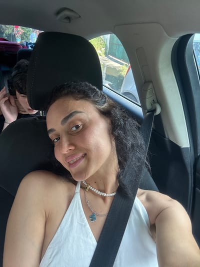 The image shows a woman in the passenger seat of a car, smiling at the camera. She has dark, wavy hair and is wearing a white dress and a pearl necklace with a blue cross pendant. A black seatbelt is across her body. In the background, a young person is visible in the back seat, with their hand near their ear. The car appears to be in motion, as indicated by the blurred background and the outdoor scenery visible through the windows, which suggests an urban or suburban environment consistent with Batumi, Georgia, possibly on a sunny day. Luggage is visible in the back seat of the car.