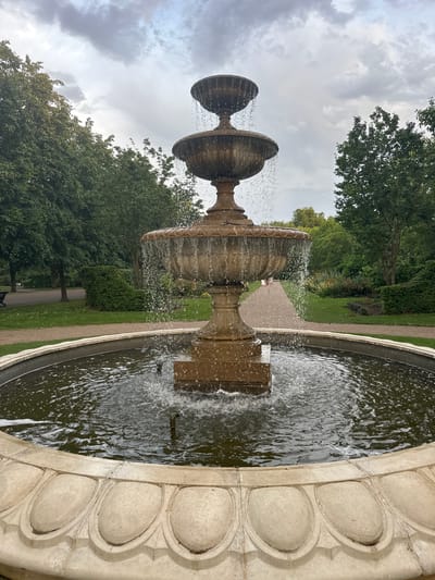 This outdoor scene shows a tiered stone fountain in a circular basin. Water is actively flowing from the fountain into the basin, creating droplets in the air. The basin has a decorative rim with oval-shaped indentations. The background features greenery, including trees and grass, suggesting a park or garden setting. A paved path is visible behind the fountain. The sky is overcast with grey clouds. It depicts a typical view of the Italian Gardens in Hyde Park, London, United Kingdom.