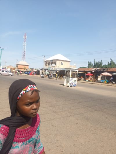 This image shows a street scene in Tumfure, Nigeria. In the foreground, a young girl wearing a black hijab and a pink and teal patterned dress is looking to her right, away from the camera. Behind her, a paved road is visible with several vehicles, including a silver car and multiple motorcycles. On the right side of the road, there are buildings and market stalls, with some vendors and customers present. A tall red and white telecommunications tower stands in the background on the left side of the image, and a clear blue sky with a few wispy clouds covers the upper portion of the scene. The overall environment appears to be a commercial area in a town or city.