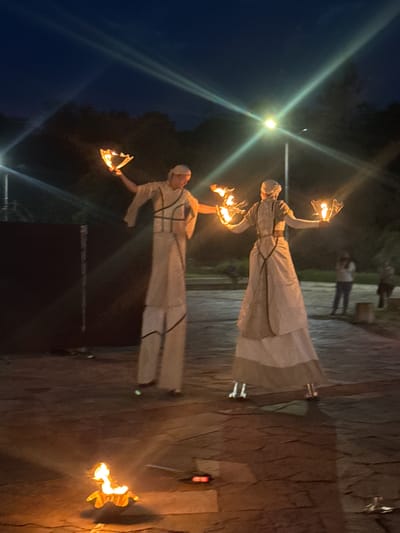 This nighttime image, captured in Plovdiv, Bulgaria, depicts an outdoor performance featuring two individuals on stilts, each manipulating fire props. The scene is illuminated by artificial light sources and the flames themselves, casting the surrounding environment into deep shadows.

In the center-left, a male performer, dressed in an off-white or light beige costume with dark, linear accents resembling straps across the torso and legs, stands on stilts. He wears a matching light-colored head covering. His right arm is extended upwards, holding a flaming object composed of multiple wicks arranged in a fan-like or semi-circular pattern, emitting bright orange and yellow flames. His left arm is bent at the elbow.

To the right of the male performer, a female performer, similarly dressed in a layered, light-colored long dress or skirt with dark straps crossing her back, also stands on stilts. She wears a light-colored head covering and faces slightly away from the camera, engaging with the performance space. She holds an identical flaming prop in each raised hand.

In the immediate foreground, positioned centrally on the paved ground, a small, low-lying object emits a significant, steady flame. The ground itself is composed of dark, irregularly shaped paving stones.

The background is predominantly dark, suggesting dense foliage or trees, with several blurred figures visible on the far right, indicating an audience or other individuals present. A prominent street lamp is situated in the upper-center, emitting a strong white-yellow light and creating distinct lens flares that traverse the image diagonally. The overall lighting is low-key, emphasizing the bright flames and the performers' illuminated costumes against the dark surroundings.