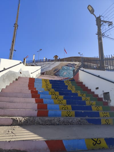 The image shows a vibrant, colorful staircase leading up a hill under a clear blue sky. The steps are painted in a repeating pattern of pink, red, blue, yellow, and green. Black decorative symbols are painted on the yellow and blue steps. The staircase appears to be part of a public space, possibly a park or a viewpoint, given the presence of a metal railing and lampposts.

In the background, a mural depicting stylized blue figures is visible on the wall of the hill. A red flag is flying from a flagpole on the upper part of the hill, and there are some buildings or structures visible at the top. The scene suggests a bright, sunny day. The location is identified as Arica, Chile. The overall impression is one of an artfully decorated outdoor public space, possibly a tourist attraction. There is no visible text in the image.