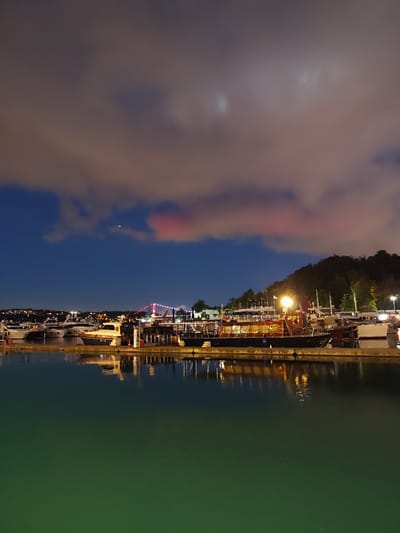 This is a nighttime, vertical, full-shot photograph taken from a waterside vantage point, looking towards a marina. The sky above is a deep blue, with scattered clouds tinged with pink and orange from artificial lights. The marina is filled with numerous boats of various sizes docked along a quay. Many of these boats are illuminated, with lights glowing from within and along their decks. The water in the foreground is a dark, greenish hue, reflecting the lights of the boats and the sky, creating a shimmering effect.
In the background, a hill is visible, covered in trees and dotted with lights from houses or buildings. A bridge with illuminated red lights spans across a body of water in the distance, possibly connecting two parts of the city. The overall mood of the photograph is serene and tranquil, evoking a sense of peace and quiet at night. The time of day is clearly night, as indicated by the dark sky and the presence of artificial lights. While no specific event is depicted, the scene captures the ambiance of a marina at night, with boats at rest and the city lights twinkling in the distance.
No people are visible in the frame. The prominent objects are the boats, the quay, the water, the bridge, and the hilly landscape with distant lights. The setting is a marina, likely in Sarıyer, Turkey, given the provided location context. There is no visible text in the image.