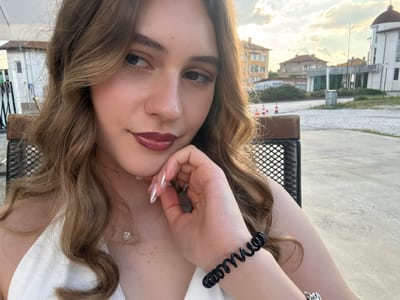 The media file is a close-up, over-the-shoulder selfie of a young woman with light brown, wavy hair. She is wearing a white top and a black spiral bracelet on her left wrist. A thin silver necklace with a heart-shaped pendant is visible around her neck. Her right hand is resting on her face, with her fingers touching her cheek and chin, showcasing manicured nails with a pearlescent finish. She has on dark lipstick.
The background is out of focus but appears to be an outdoor setting with buildings and a cloudy sky, suggesting it is daytime. The orientation of the image is rotated. Based on the provided location context, the scene is likely Panagyurishte, Bulgaria. The overall mood conveyed by the image is casual and relaxed. No visible text is present in the media file.