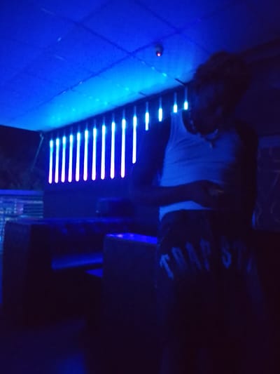 A person wearing a white tank top and dark pants with "TRAP STAR" printed on them stands indoors. The location is dimly lit with a bluish hue. A row of bright vertical lights is visible in the background, likely illuminated by neon lights. Furniture is also present, suggesting the scene is set in a bar. The presence of the person looking at their phone, the lights, and furniture indicate that the subject is at leisure. There is no visual confirmation for the exact location of Narayi, Nigeria.