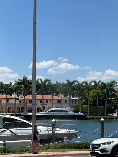 In Miami Beach, Florida, USA, a person stands on the sidewalk next to a body of water, looking out at several boats and a large house. The person is wearing a dark t-shirt and patterned shorts. In the water, there are at least two yachts docked. Behind the yachts, a multi-story building with a terracotta roof and arched windows is visible, surrounded by palm trees. The sky is blue with scattered white clouds. A white SUV is parked on the right side of the image.