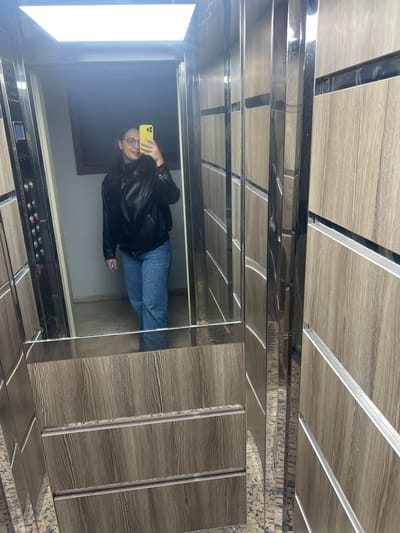 A person in a black jacket and blue jeans takes a selfie in a mirrored elevator. The phone, held in the left hand, is yellow. The elevator has a mirrored wall and a control panel visible. Above, the ceiling panels feature a light wood grain pattern with thin silver stripes. The elevator's side walls are covered in matching wooden panels with silver edges.