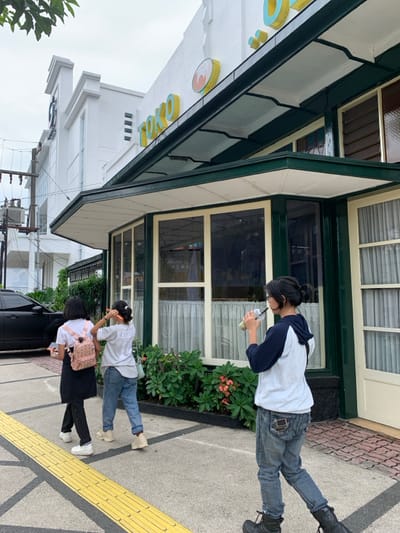 Three young women stand outside a storefront in Malang, Indonesia, at the coordinates -7.981 latitude and 112.63 longitude. The building is white and has a green awning. The awning reads "TOKO" in yellow letters. One woman is wearing a white long-sleeve shirt and blue jeans. She has her hair pulled back in a ponytail and is looking down at the ground as she drinks from a clear plastic cup with a straw. The other two women are wearing black long-sleeve shirts and black pants. One of them is wearing a pink backpack and the other has a green backpack. The woman with the pink backpack is holding a black smartphone. The woman with the green backpack has her hair pulled back in a ponytail. They are both looking at the woman drinking the beverage. The ground in front of the women is gray concrete with yellow lines painted on it. The building's exterior is made of white brick. There are green bushes in front of the building. A black car is parked in the background, partially visible.
