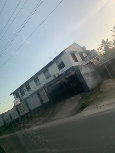 A long building with many windows is visible in the image. A road runs parallel to it. The photograph was likely taken from inside a vehicle. The sky is overcast. There are electrical wires in the foreground. Palm trees are visible in the background. Based on the architecture and surroundings, this image is potentially from Dar es-Salaam, Tanzania.