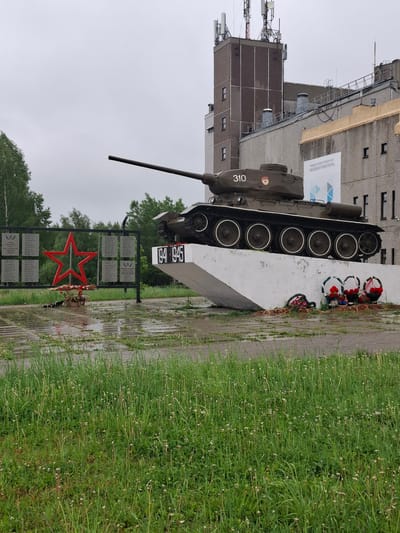The image shows a T-34 tank monument in Марковский, Russia, commemorating Soviet soldiers in World War II. The tank is mounted on a concrete pedestal with the years 1941-1945 inscribed on it. Wreaths are laid on top of the monument. A black metal sign with a red star and names is erected nearby. The surrounding area is green with grass and trees. Buildings, some tall, can be seen in the background. The weather appears overcast and possibly rainy, as the ground looks wet.