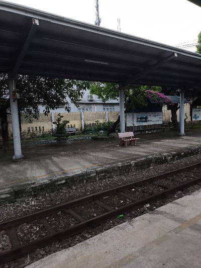 This image depicts a deserted train station platform in Thành phố Quảng Ngãi, Vietnam, under an overcast sky. In the foreground, two parallel railway tracks with dark sleepers and grey ballast run horizontally across the bottom, separated from the platform by a weathered concrete edge. A small green piece of litter is visible on the ballast between the tracks.
The midground features a concrete platform covered by a dark grey corrugated metal roof supported by light grey concrete pillars. On the platform, a single reddish-brown wooden bench with a backrest faces away from the camera, positioned to the right of a grey concrete pedestal holding a small potted plant. Behind these, a longer, unpainted wooden bench without a backrest is visible. Further back, a vibrant bougainvillea plant with purple flowers drapes over a small structure or tree. To the far right, a large white information board with text and images is affixed to a wall or structure.
In the background, a light yellow concrete wall with a patterned texture separates the platform area from a multi-story building. This building features light grey walls and numerous rectangular windows, with several external air conditioning units mounted on its facade. Scattered green trees and shrubs are visible both behind the wall and within the station's immediate surroundings. Above the building, multiple communication towers or antennas are visible against the grey sky. No individuals or active interactions are present in the scene.