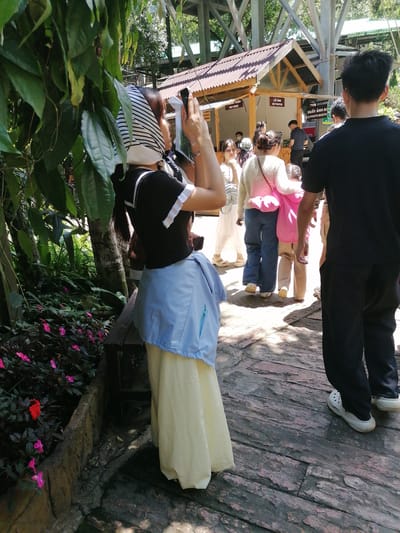 The image depicts a scene at an outdoor attraction in Da Lat, Vietnam, likely a tourist spot given the presence of a photo booth and signage in Vietnamese and English. The foreground is dominated by lush green foliage and pink flowers, suggesting a garden or natural setting. A woman in a black top, white and black striped headscarf, and a light blue and yellow dress is seen from the back, holding up a phone to take a photo. To her right, a man in a black t-shirt and black pants is walking away from the camera. Behind them, a group of people, including children, are milling around a small wooden structure with a brown roof that appears to be a service area or shop. Signs on the structure indicate "PAY HERE" and "GET PHOTOS." The ground is paved with rough, reddish-brown stones. The overall atmosphere is bright and sunny, with dappled sunlight filtering through the trees and structures.