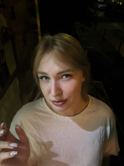 The image shows a young woman with blonde hair and light-colored eyes, wearing a white t-shirt. She has on a light makeup, with emphasis on her eyes and lips. Her nails are long and painted with a white design. She is holding up her left hand towards the camera. The background is dark and appears to be an outdoor urban setting at night, possibly in Khimki, Russia, with some indistinct structures and a street visible.