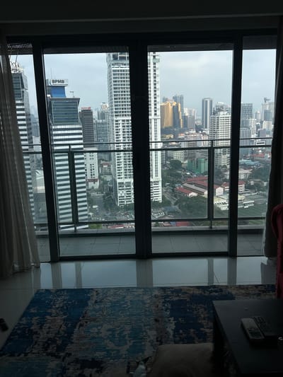 The image shows a view from a high-rise building in Kuala Lumpur, Malaysia, looking out over a dense cityscape. The perspective is from an interior room, with sheer curtains on the left and a portion of a dark side table and a patterned rug visible in the foreground. The main focus is the expansive urban landscape visible through a large glass window, divided by vertical mullions.
The cityscape is characterized by numerous skyscrapers of varying heights and architectural styles. Tall, slender buildings with many windows dominate the scene, interspersed with shorter, blockier structures. Some buildings feature distinctive designs, such as the white, patterned facade of a prominent skyscraper on the left side of the view. Another notable building bears a blue sign with "BPMB" and "Malaysia Development Bank" written in white lettering. In the mid-ground, a cluster of golden and grey buildings stands out. Lower-rise buildings with red roofs are visible further in the distance, along with roads and what appears to be a sports field or park area. The sky is overcast, suggesting a cloudy day. The overall impression is one of a bustling, modern metropolis.