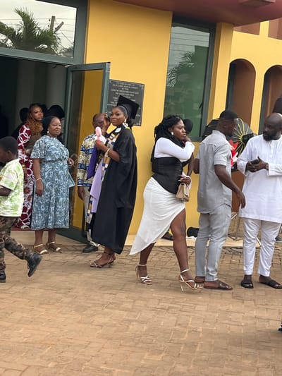 The image depicts an outdoor gathering of approximately eight individuals in front of the C.E.M World Missionary Headquarters in Madina, Ghana. The building has a yellow facade with green-tinted glass doors and windows, and a black plaque inscribed with "TO THE GLORY OF OUR LORD JESUS CHRIST & IN TRUST OF C.E.M WORLD MISSIONARY HEADQUARTERS" is affixed to the wall. The ground is paved with reddish-brown interlocking bricks.
From left to right, a young boy in a light green patterned shirt and camouflage pants walks toward the left foreground. An older woman in a light blue patterned dress and gold sandals stands near a glass doorway, observing the scene. Behind her, an older man in a colorful Kente-print shirt is partially visible. Central to the frame is a young woman, presumably a graduate, wearing a black graduation gown, mortarboard cap, and a purple and gold Kente-patterned stole; she holds a pink handheld fan to her left cheek and wears silver high-heeled sandals. To her right, another young woman with dreadlocks wears a white long-sleeved top, a black corset, a white asymmetrical skirt adorned with small white pearls, and gold high-heeled sandals, carrying a light-colored handbag. Next to her, a man in a matching grey short-sleeved shirt and trousers stands with his back partially to the camera. Further right, an older man with a beard, dressed in a white traditional Ghanaian long-sleeved outfit and dark sandals, stands with clasped hands, looking towards the left. Additional individuals are partially visible near the doorway and in the background. The scene is illuminated by natural daylight, and a palm tree is reflected in one of the building's glass panels.