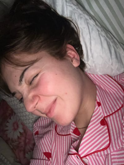 A woman is sleeping on a pillow with a striped case. She is wearing pink and white striped pajamas with red accents. Her eyes are closed, and her face is relaxed. Her dark hair is slightly disheveled. Part of a floral patterned blanket is visible beneath her. The scene is dimly lit.