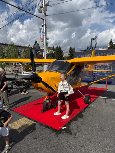 A yellow propeller airplane is parked on a red carpet in front of a digital display screen. A young girl with braided hair, wearing a white patterned shirt and black shorts, stands on the carpet next to the airplane. Her arms are down and she holds a black umbrella in her left hand. In the background, a statue of a man is visible, along with trees and buildings. The sky is partly cloudy. The image is an outdoor scene, possibly a public event or exhibition.