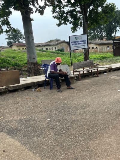 A person wearing a pink and yellow head covering, a white shirt, and purple vest is sitting on a blue chair by a road in Nigeria. They are holding a phone in their hand, appearing to be engaged with it. Beside the person, there is a white sign with the text "ANTI-CORRUPTION & TRANSPARENCY UNIT (ACTU) UNIVERSITY OF JOS" and contact information. The scene appears to be outdoors, with a tree on the left and buildings in the background. The road is made of asphalt, and there are some benches and a concrete drain visible. The weather appears to be overcast, suggesting it might be morning or late afternoon. The overall setting suggests an institutional or educational environment, given the university sign.