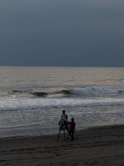 A forensic examination of the image reveals a beach scene in Belalang, Indonesia, under an overcast, dark gray sky. The dark, wet sand occupies the foreground and midground, leading to a dark ocean with multiple white-capped waves breaking near the shore. Two adult males and a white horse are present. One male, wearing a light-colored sleeveless top and dark sunglasses, is mounted on the horse. The second male, dressed in a red short-sleeved shirt and dark bottoms, walks directly beside the horse, holding its bridle, indicating he is leading the animal. Both individuals and the horse are positioned near the water's edge and are moving generally parallel to the shoreline. The overall lighting is dim, consistent with dusk or heavily overcast conditions.