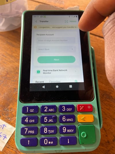 A close-up, top-down, angled outdoor shot shows a green and black point-of-sale (POS) device on a wooden surface. The device has a touchscreen displaying a transfer interface with fields for "Recipient Account" and "Select Bank," and a "Next" button. A finger is obscuring the upper right corner of the screen. The device is surrounded by a wooden surface that appears to be a table. To the left of the POS device, a torn piece of paper with handwritten text is visible. The overall scene is likely in Mararaba, Nigeria, given the context.