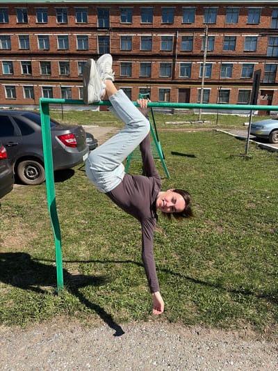 A woman is posed horizontally on a green metal jungle gym, with her body angled toward the camera. She wears a dark long-sleeved top, light blue jeans, and white sneakers. Her left leg is bent and her foot rests on the gym while her right leg is straight. Her left arm holds onto the gym, and her right arm is extended outward. She is smiling at the camera.

Behind her, a large brick building with multiple windows is visible. A car is partially visible beneath the jungle gym and another car is partially visible on the upper-left. The ground consists of grass and gravel. The scene is located in Neftekamsk, Russia.