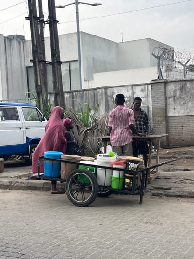 In Itirin, Nigeria, an outdoor scene shows a street side vending cart. Two men stand at the cart, one in a pink striped shirt and the other in a plaid shirt, appearing to be interacting at the cart’s counter. The cart is loaded with multiple buckets and containers. A woman in a pink hijab stands near the cart beside a blue and white vehicle. Overhead, power lines run along a building, and a fence topped with barbed wire is visible. The surrounding environment consists of buildings and a paved street.