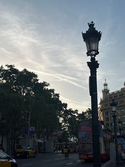 This image captures an outdoor urban scene, likely taken in Barcelona, Spain, given the presence of a bus with Catalan text. A prominent street lamp with an ornate, dark metal design stands in the foreground, silhouetted against a hazy sky. The sky itself features wispy, white clouds, suggesting either early morning or late afternoon light.

Behind the street lamp, a multi-story building with arched windows and classical architectural details lines the street. To the right, a red and colorful double-decker bus is visible, adorned with text that appears to be advertising "Poble Espanyol" and describes it as a place where "History Meets Culture and Leisure." Further down the street, yellow taxis are parked, and lush green trees line the sidewalk, partially obscuring the view of other buildings and streetlights. The overall impression is one of a bustling city street during a calm moment, perhaps before the height of activity for the day. The light and the presence of the bus suggest it could be a tourist area. There are no discernible people in the foreground, and the focus is on the streetscape and architectural elements.