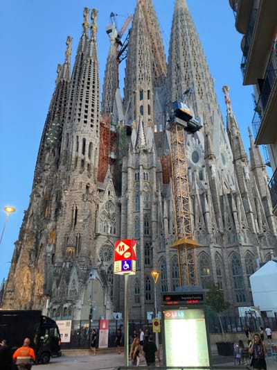 The image shows the Sagrada Familia in Barcelona, Spain, under construction. Several towering spires of the basilica, constructed from stone and adorned with intricate carvings, dominate the frame against a clear blue sky. Cranes are actively working on the upper sections of the building, with scaffolding and construction materials visible.

In the foreground, a street scene unfolds. A dark-colored van is parked on the left. To the right of the van, a signpost displays the Barcelona Metro logo, indicating a subway station, along with directional arrows and information about lines L2 and L5. Above this, another sign reads "Sagrada Familia". A lamppost with an orange light fixture stands to the left of the basilica.

Several individuals are present on the street. Some are walking, while others appear to be standing or conversing. One person in the lower left corner is wearing an orange safety vest. The overall atmosphere suggests an active urban environment where tourism and ongoing construction coexist. The lighting indicates it is either late afternoon or early evening.