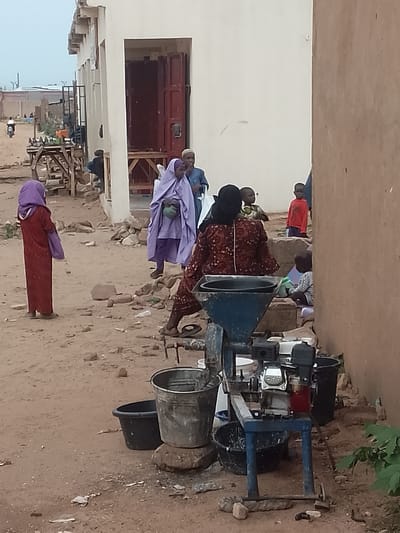 The image depicts an outdoor scene in the city of Gombe, Nigeria, characterized by sandy ground, scattered rocks, and simple building structures. In the foreground, a young girl wearing a long red dress and a purple headscarf stands facing away from the camera, holding a purple bag. To her right, a woman in a light purple hijab and traditional attire stands with her back slightly turned, carrying a green bag. A man in a blue traditional outfit and a cap is partially visible behind her.
Further back, a group of children are present. One child in a red shirt stands looking towards the camera. Another child wearing a striped shirt is seated on the ground. Behind this group, a woman in a patterned brown dress is seated near a blue metal machine that appears to be a grinder or mill, with buckets placed around it.
The environment suggests a semi-urban or rural setting with rudimentary infrastructure. The buildings are simple, one appearing to be a pale-colored structure with an open doorway, and another a darker, earthy-toned wall on the right. In the background, a wooden cart with various items is visible, and beyond that, more buildings and a person on a motorbike. The overall impression is a snapshot of daily life in a community.