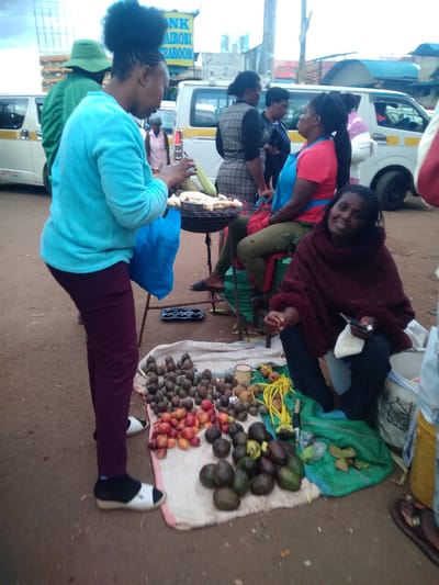 The image captures an outdoor market scene in Nyeri, Kenya, under daylight. In the foreground, a woman wearing a teal long-sleeved top, maroon trousers, and black and white sandals stands, holding a blue bag in her left hand while preparing an item, possibly corn, on a metal charcoal grill. Her hair is styled in a braided updo. To her right, a second woman is seated on a red plastic stool, wearing a red t-shirt and a blue apron. To the immediate right, a third woman is seated directly on the reddish-brown ground, wrapped in a dark maroon, fuzzy poncho over dark clothing. She holds a mobile phone in her left hand and a small white packet in her right, smiling towards the viewer.

On the ground in front of the seated woman, a light-colored cloth displays various types of produce: large piles of avocados, smaller round red and orange fruits, and numerous small, dark, round fruits. A yellow metal mug, a knife, and yellow string-like objects are also present on the cloth, which is partially covered by a green fabric. In the background, several white passenger vans with yellow stripes, consistent with Kenyan matatus, are visible. A yellow sign with partially visible text, "NK NAIROBI TEAROOM," hangs above the vehicles. The ground is unpaved, composed of reddish-brown dirt and gravel. Multiple other individuals are present in the mid-ground and background, indicating a busy urban street or market environment.