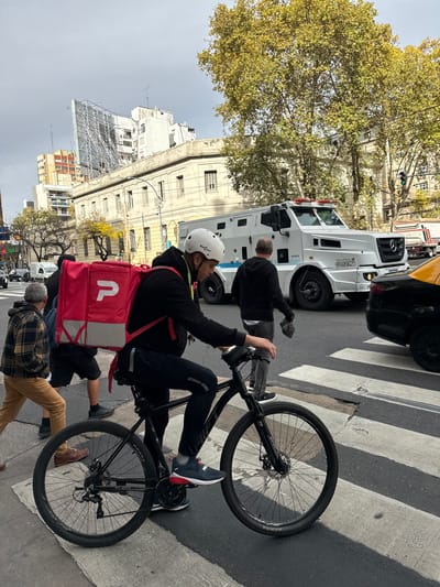The image shows a street scene in Buenos Aires, Argentina. A cyclist with a white helmet and a red delivery backpack is riding a bicycle on a zebra crossing. An armored truck, a yellow cab, and other vehicles are visible in the background. A man is walking past the armored truck. There are several other pedestrians. Buildings and trees line the street.