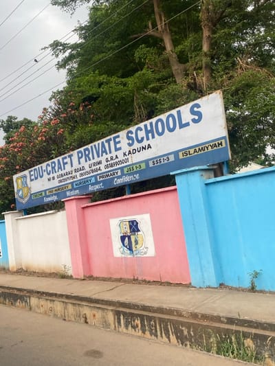 The image depicts the exterior of "EDU-CRAFT PRIVATE SCHOOLS" located at NO 18 GOBARAU ROAD, U/RIMI G.R.A KADUNA, Nigeria. This scene is presented in the context of the city of Rido, Nigeria.
A large, weathered rectangular signboard with a white background and prominent blue lettering displays the school's name, address, contact numbers (08059797549, 0806578684, 08189905001), and lists educational stages: CRECHE, PRE-NURSERY, NURSERY, PRIMARY, JSS 1-3, SSS 1-3, and ISLAMIYYAH. Below these, values such as Knowledge, Wisdom, Respect, and Confidence are noted. A school crest, a shield featuring an open book and crossed implements, is present on the left side of the sign.
Below the signboard, a low perimeter wall runs parallel to the roadside. This wall is sectioned and painted in three distinct colors: white on the left, pink in the center, and light blue on the right. A faded rendition of the school crest is painted on the pink section of the wall. The base of the wall and the narrow concrete sidewalk in front of it show signs of dirt and sparse vegetation. In the immediate foreground, a concrete curb and gutter system runs the length of the wall, appearing discolored and worn.
The upper portion of the image reveals dense green foliage from trees, with some flowering plants exhibiting reddish-pink blossoms behind the left side of the sign. Utility wires are visible traversing the upper left quadrant. The sky is overcast. No individuals are present in the depicted scene.