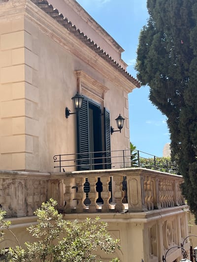 The image shows a close-up view of a building facade and balcony in Taormina, Italy, under a bright blue sky with scattered clouds. The building is constructed with light-colored stucco and features a terracotta tiled roof with a decorative cornice. A pale yellow wall is adorned with fluted pilasters. Two black wall-mounted lanterns illuminate a partially open window with dark green shutters. A stone balcony with ornate balusters and a wrought-iron railing overlooks a lush green tree that dominates the right side of the frame. In the foreground, a small tree with light green leaves adds to the Mediterranean ambiance.