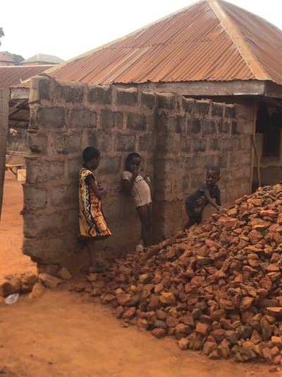 The image portrays an outdoor scene in Nsugbe, Nigeria, featuring three children adjacent to a building and a large pile of rocks. The ground consists of reddish-brown dirt. A building with an unrendered concrete block wall is situated on the right, topped by a weathered, reddish-brown corrugated metal roof. Another structure with a reddish roof is visible in the background, further left.

Three individuals identified as children are present. On the left, a child wearing a multi-colored patterned dress stands on the dirt ground, facing right, with her left hand raised towards her mouth. In the center, a child in a light-colored t-shirt leans against the concrete block wall, holding a small, light-colored object to their mouth. To the right, a third child, dressed in a dark t-shirt, stands near and partially on a large pile of reddish-brown rocks, with one hand resting on the rocks. A piece of white plastic debris is visible on the ground in the lower-left quadrant.