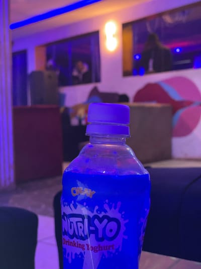 The image shows a close-up of a blue plastic bottle containing "NUTRI-YO Drinking Yoghurt." The bottle is partially filled with a light-colored liquid, and condensation is visible on the exterior, suggesting it is chilled. The bottle has a white cap with ridges. Text on the label reads "NUTRI-YO Drinking Yoghurt" and "CRAZY" in a stylized font. The background is blurred but appears to be an indoor setting with purple and blue lighting, possibly a restaurant or lounge. There are hints of artwork or posters on the wall in the background, illuminated by the ambient light. The overall scene is dimly lit, with the blue bottle being the primary illuminated subject. The location is noted as Durumi, Nigeria.