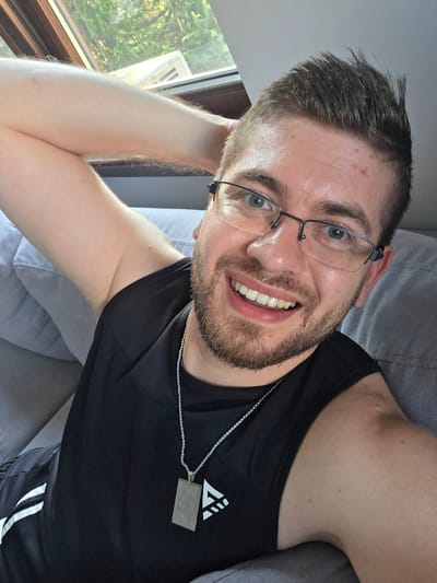 A young adult male is captured in a close-up, diagonal selfie, smiling broadly while lying on a light gray sofa or cushion. He is positioned with his head towards the bottom right of the frame, looking upwards directly at the camera. He has short brown hair, a well-trimmed beard and mustache, and fair skin, with visible blue-green eyes behind clear-rimmed glasses. His mouth is open in a wide, relaxed smile, showcasing white teeth.
He is wearing a black sleeveless athletic top, on which a silver chain necklace with a rectangular, metallic grey pendant is visible. The top features a white triangular graphic with a prominent "W" letter on the left side of his chest. In the upper left corner of the image, black athletic shorts with white stripes are partially visible. One of his arms is bent and raised, resting behind his head, while the other is mostly out of frame.
The background shows bright natural light streaming in from a window in the upper right, revealing lush green foliage and trees outside, suggesting a sunny daytime setting. The overall mood is relaxed and happy. The location is indoors, likely a residence in Yagodovo, Bulgaria.
