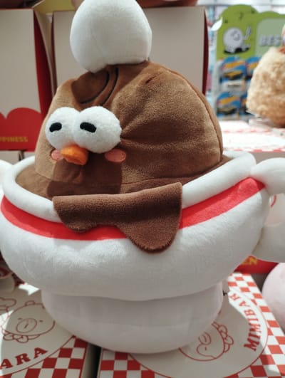 The image displays a close-up of a plush toy designed to resemble a dessert, possibly a cup of ice cream or a similar treat. The toy is predominantly white and brown, with a large white sphere at the top, resembling a scoop of cream or ice cream. The main body of the toy is brown, textured like soft fabric, and shaped like a bowl or cup.
The plush toy has distinct facial features. Two large, white circular eyes with black pupils are positioned prominently on the brown body. A small, orange, carrot-shaped nose sits below the eyes, giving the toy a cute, cartoonish appearance. Pink circles are placed on either side of the nose, possibly representing cheeks or blush. A brown, curved piece of fabric drapes over the front of the brown body, resembling a flap or a blanket.
The toy is sitting in a white bowl-like structure, which is encircled by a red and white striped band. The background is blurred, but suggests an indoor retail environment. To the left, a red box with white text is partially visible. Part of the text "HAPPINESS" is legible. To the right, shelves with various items are out of focus, including what appear to be packaged goods, possibly toys or collectibles. Some of these items have green packaging with cartoon illustrations.
A round, white sticker with red text and an illustration of a cartoon character is visible on the surface beneath the plush toy. The text on this sticker includes "MIMIBAR" and "ARA". The overall presentation suggests a display of merchandise in a store. The lighting appears to be artificial, typical of an indoor setting. No specific time of day or weather conditions are discernible. The emotion conveyed by the toy is cheerful and whimsical. The location context provided states Nha Trang, Vietnam, which hints at the retail setting potentially being in this city.