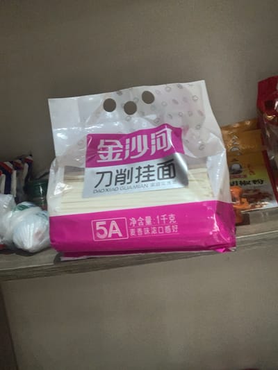 In Abuja, Nigeria, a package of noodles is displayed on a shelf. The package is predominantly pink and white and features Chinese characters and the "5A" logo. The noodles inside are thin and white. To the left of the noodle package, several white plastic-wrapped items are visible. In the background, a yellow package with red and orange lettering is partially obscured. The lighting suggests an indoor environment.