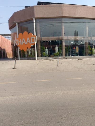 The image shows a storefront for "Khaadi" in Peshawar City Tehsil, Pakistan. The building features a prominent orange sign with the store's name. The facade has large glass windows, reflecting the sky and possibly buildings opposite. Inside, mannequins are visible, displaying clothing. Several potted plants are placed strategically. The area in front of the store appears to be a paved walkway or parking area, with black stanchions and yellow road markings visible. The sky is clear and blue. The overall scene suggests a commercial establishment in an urban setting.