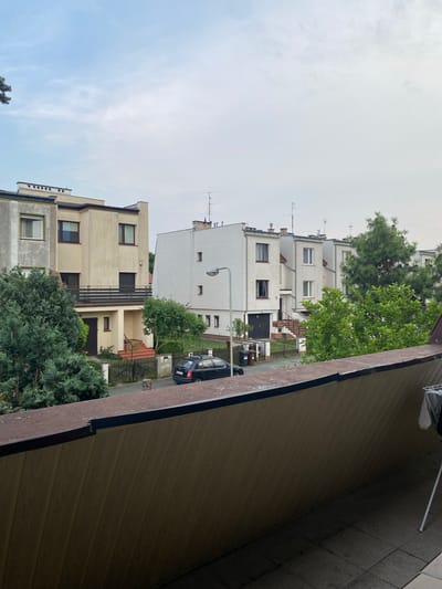 A medium-shot, eye-level, outdoor photograph captures a residential street scene in Wrocław, Poland, under a cloudy sky. In the foreground, a wooden-paneled balcony railing is visible, with a clothes drying rack on the right side. Beyond the railing, a row of multi-story buildings lines the street, interspersed with trees and foliage. The buildings appear to be terraced or attached townhouses, with a mix of architectural styles. Some have light-colored facades, while others are darker. A black car is parked on the street in front of one of the houses, and a wooden fence separates the properties from the road. The overall atmosphere is one of a quiet, suburban neighborhood.