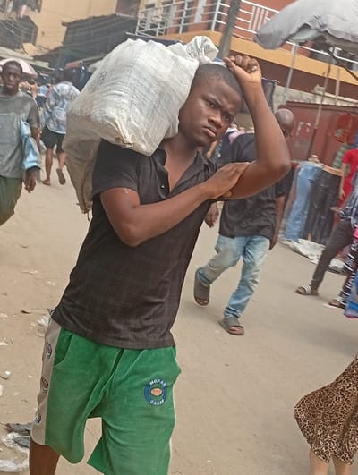 The image portrays an outdoor street or market scene in Fegge, Nigeria, under daylight conditions. A young, dark-skinned male, approximately 18-25 years old, is the central figure, facing slightly left of the viewer. He is dressed in a black short-sleeved polo shirt and green and beige shorts, the latter bearing a circular "MOPAR GARAGE" logo on the left thigh. He carries a large, white, woven sack over his right shoulder. His left hand is raised, resting on the back of his head/neck, while his right forearm is bent across his body. The ground is a light-colored, unpaved surface with some debris. In the background, multiple pedestrians are visible, along with market stalls or buildings constructed from materials like corrugated metal. An open grey market umbrella is partially visible on the right side. Individuals in the background include a person in a light shirt on the left and another in blue jeans and sandals on the right.